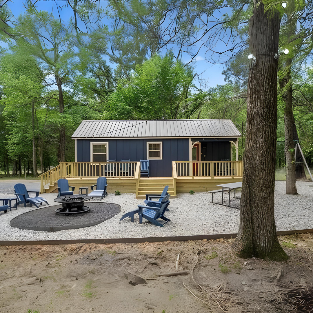 Social posts starting 7924 27 - Lelands Modular - Lelands Modular Blue cabin with a metal roof on a wooden deck, surrounded by trees, and blue Adirondack chairs around a fire pit.