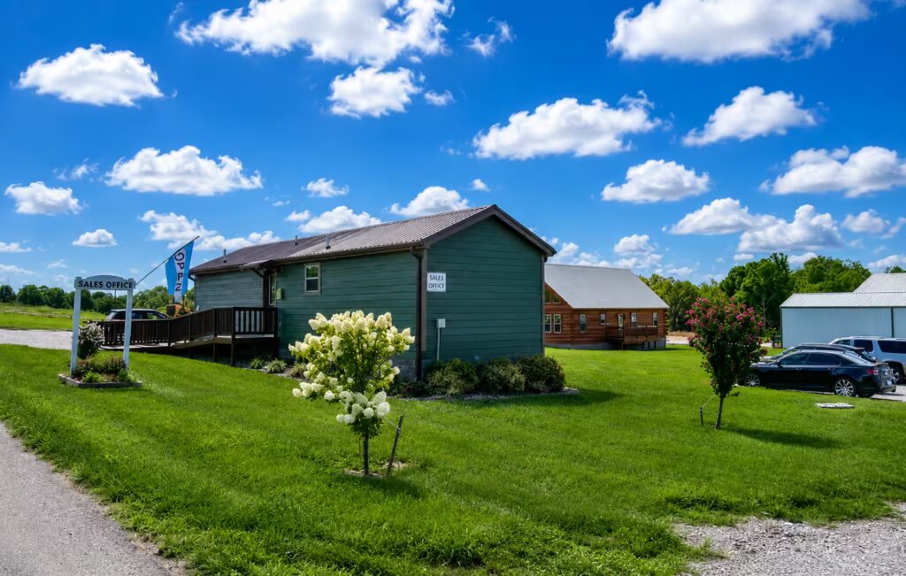 Green wooden sales office on a sunny day; sign says 'Sales Office' near the entrance beneath a blue sky with white clouds.