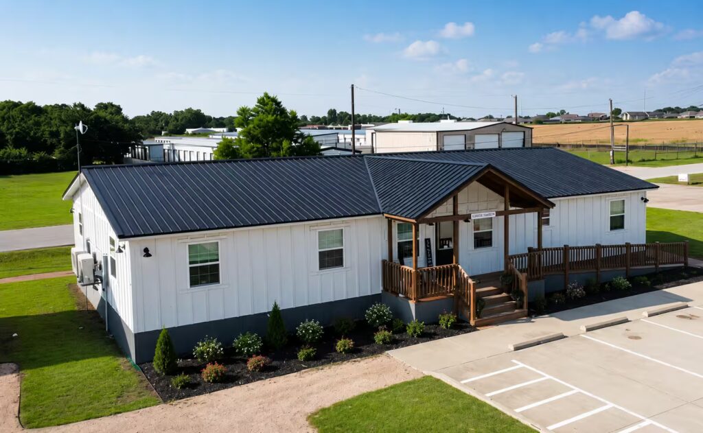 White single-story commercial building with a dark metal roof and wooden front porch, parking lot in foreground.